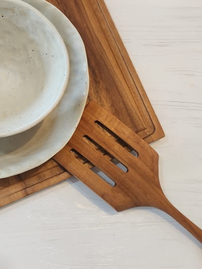 The image shows a close-up, overhead view of kitchenware arranged on a white, textured surface. The primary objects are two off-white ceramic bowls, one nested inside the other, and a wooden spatula. The spatula is made of a warm-toned wood and has several rectangular slots in its wider end, suggesting it's a serving or cooking utensil. It lies diagonally across the frame, with its handle extending towards the top right corner.
Beneath the spatula, partially visible, is a rectangular wooden cutting board. The cutting board has a subtle groove around its perimeter. The ceramic bowls are positioned in the lower left quadrant of the image. They have a speckled, handmade appearance, with visible texture and small dark flecks against the off-white glaze.
The white surface beneath the objects appears to be painted wood, with visible brushstrokes that create a slightly irregular texture. The lighting is soft and diffused, casting no harsh shadows, and suggests an indoor setting, possibly a kitchen or dining area. There is no indication of a specific time of day, weather, or any discernible emotions. No text is visible in the image. The overall aesthetic is rustic and simple, highlighting the natural materials of the wood and ceramic.