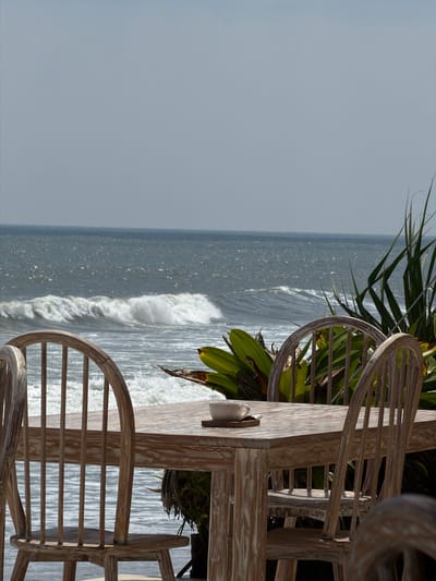 The image displays an outdoor dining or lounge area situated on a coastline in Belalang, Indonesia. A rectangular wooden table with a distressed light finish occupies the central foreground, surrounded by several matching wooden chairs featuring spindle backs. On the table, a white ceramic cup is placed on a small, dark wooden coaster, next to a small, dark rectangular object. Directly behind the table, a dense growth of green tropical foliage, appearing as large-leafed plants, provides a natural barrier. Beyond the foliage, the ocean extends to the horizon, characterized by active waves with visible white crests breaking across the dark blue-grey water. The sky above the ocean is a clear, light blue-grey, indicative of an overcast or hazy condition. No individuals are present in the scene.