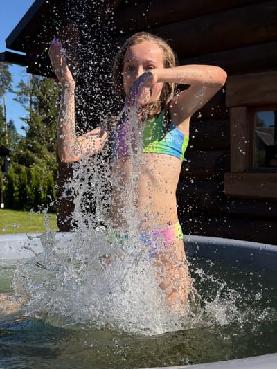 A young girl with blonde hair is captured mid-air, splashing into a body of water, likely a pool or hot tub. She is wearing a colorful bikini with a tie-dye pattern featuring shades of green, blue, pink, and purple. Her face shows a mixture of exhilaration and surprise, with her eyes wide open and her mouth slightly agape. Water droplets fly around her, frozen in motion by the camera's shutter speed, creating a dynamic and energetic effect.
The background suggests an outdoor setting on a sunny day. To the left, a rustic wooden structure, possibly a cabin or sauna, is visible. Trees and a bright blue sky can be seen in the distance, indicating it's likely daytime and the weather is fair. The overall impression is one of carefree summer fun and a moment of pure joy. No visible text is present in the image.