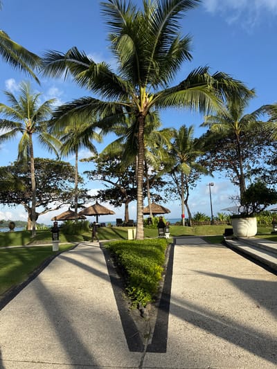 The image depicts an outdoor scene in Jimbaran, Indonesia. The foreground features a paved walkway with a dark border, lined with green shrubs and palm trees casting shadows across the path. In the midground, there are thatched-roof structures resembling beach umbrellas or small huts. The background reveals a glimpse of the ocean and a blue sky with scattered clouds. No individuals are clearly visible in the scene. The environment suggests a tropical or coastal resort area.
