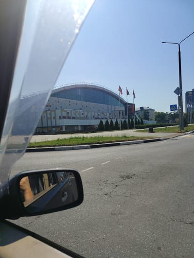 The image, taken from inside a vehicle, shows the Salihorsk Ice Palace in Salihorsk, Belarus, on a clear, sunny day. The modern, curved building features a facade with a grid of colorful rectangular panels in yellow, orange, and brown. Three flags, one red and two red and green, fly in front of the building. A tall streetlight stands to the right of the building, along with pedestrian crossing signs. The foreground is dominated by a cracked asphalt road and a car's side mirror, which reflects a person's arm and hand. In the background, other buildings and trees are visible.