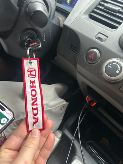 The image displays the interior of a vehicle, focusing on the driver's side dashboard and center console. A human hand, with fair skin, holds a white rectangular fabric keychain featuring "HONDA" in red lettering and the Honda logo, which is attached to a metal key ring. The key ring is connected to a black key fob, which is inserted into the ignition switch located on the steering column. The dashboard, made of gray plastic, shows an air vent, a red triangular hazard light button, and a rotary fan speed control knob. Below these controls, a black car charger with a red illuminated ring is plugged into an accessory power outlet, with two black charging cables extending from it. A partial view of a smartphone screen is visible in the lower-left, indicating a "5G" network connection and what appears to be a voice assistant interface. A light-colored piece of clothing or fabric is visible below the hand. The specific location within Medford, United States, is not discernible from the image.
