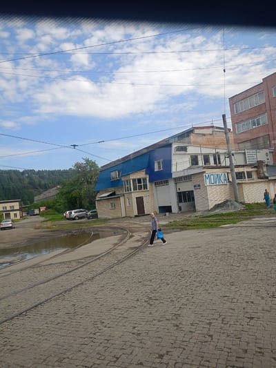 The image depicts a street scene in Zlatoust, Russia, with a focus on a car wash facility. In the foreground, tram tracks are laid on a cobblestone surface, with a puddle of water to the left. A woman is walking across the tram tracks, carrying a blue plastic bag. Behind her, a building complex includes a car wash with "МОЙКА" (Car Wash) written on its facade. Several cars are parked in the vicinity, and a hill covered in trees forms the backdrop. The sky is partly cloudy.