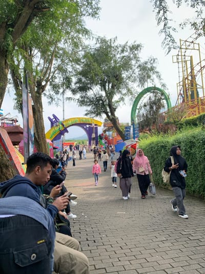 The image shows a group of people walking on a brick pathway at Jawa Timur Park 1 in Batu, Indonesia, located at latitude: -7.885, longitude: 112.524. The park entrance is visible in the background, adorned with colorful arches and signs. A young girl wearing a pink shirt walks towards the entrance, while several other people walk in the opposite direction. A group of four people are sitting on a bench along the pathway, all looking down at their mobile phones. To the right of the pathway is a green bush, and beyond that, a roller coaster track and a large Ferris wheel can be seen in the distance. The sky is overcast and gray.