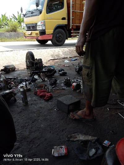 The image shows a scene in Kecamatan Haurgeulis, Indonesia (latitude: -6.414, longitude: 107.953). A person is standing next to a motorcycle that has been partially disassembled. The motorcycle is lying on its side, with its engine parts scattered around. The person is wearing sandals and shorts. They are looking down at the motorcycle. There are several tools and pieces of the motorcycle lying on the ground. In the background, a yellow truck is parked. The scene is set in a parking lot, with a dirt surface and a small amount of vegetation in the background. The photo was taken on April 13th, 2023, at 2:06 PM. The person appears to be inspecting the engine. There is a red cloth, a black box, and a spray can on the ground next to the motorcycle. The person is holding a cigarette in their right hand. The image was taken with a VIVO Y19s camera.