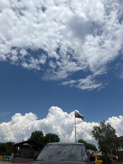 The image depicts a scene in Ravno Pole, Bulgaria, on a bright, partly cloudy day. The sky is a vibrant blue, filled with large, puffy cumulus clouds that dominate the upper portion of the frame. A Bulgarian flag, horizontally striped in white, green, and red, flies from a tall pole on the right side of the image. In the foreground, the hood and windshield of a dark-colored vehicle are visible. Behind the vehicle and to the left, a small, open-air structure with a dark-tiled roof is present. A sign with Bulgarian text and an emblem is mounted on the structure. Several trees, including a large birch tree with delicate, drooping branches on the right, are scattered throughout the scene. In the distance, to the left of the structure, two yellow and green bins are visible. A small dark shape, possibly a bird, is in the sky in the upper left quadrant. Further to the right, a bright yellow vehicle is parked. The overall atmosphere is one of a sunny day in a semi-rural or small town setting.