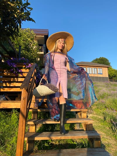 A young woman with blonde hair, wearing a wide-brimmed straw hat adorned with flowers, a light purple dress, a sheer iridescent cape, and black boots, stands on a wooden staircase. She is holding a woven basket filled with lavender. The staircase is surrounded by lush green foliage and purple flowers. In the background, a wooden building with several windows sits on a hill overlooking a field of lavender. The sky is clear and blue. The scene appears to be set in a rural area, possibly a lavender farm, during the daytime, with the sun casting long shadows. The location is Verkhneye Uchdere, Russia.
