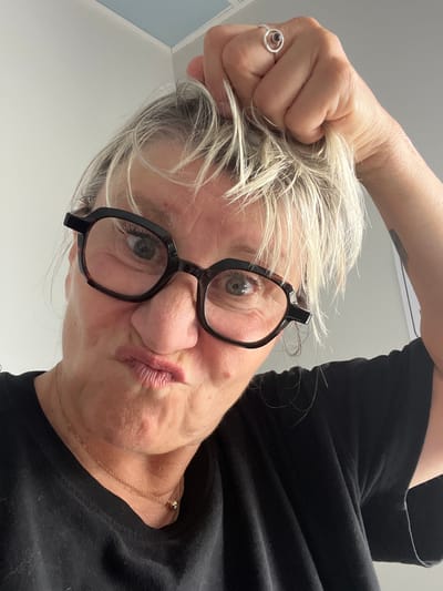 The image shows a close-up, waist-up, selfie-style photo of a woman with blonde hair and glasses. She is pulling a section of her hair with her right hand, which also wears a ring with a dark stone. Her mouth is pursed, and she is looking directly at the camera. She is wearing a black t-shirt and a delicate gold necklace. The background is a plain, light-colored wall with a sliver of a blue ceiling visible at the top. There is no indication of the image being taken in Saint-Galmier, France.