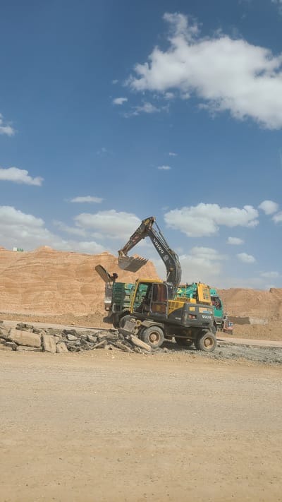 The image captures a construction or road work scene under a bright, clear blue sky with scattered white cumulus clouds, in what appears to be an arid, mountainous region of Kuchlak, Pakistan.

The primary subject is a large, yellow and dark grey Volvo wheeled excavator, specifically identified by the "EW140B" model number visible on its side. The excavator is positioned on a dirt road, its articulated digging arm and bucket extended downwards and towards the left, indicating it is actively engaged in scooping or breaking ground. Loose rocks and construction debris are scattered around the base of the machine.

Behind the main excavator, several other heavy vehicles are partially visible. Another yellow Volvo machine, possibly a different type of excavator or a front-end loader, is parked alongside it. Further back, a green dump truck or similar heavy-duty vehicle is also present. These machines are situated next to a distinct, reddish-brown geological formation, characterized by layered rock and earth, which forms a significant part of the landscape.

The ground consists of a wide dirt track or unpaved road, suggesting ongoing road construction or maintenance. The bright natural lighting indicates it is daytime, likely midday, with strong sunlight illuminating the scene. The overall setting points to an outdoor industrial operation in a rugged, dry environment. The prominent "VOLVO" text is visible on the excavator's arm and side, and also on the machinery directly behind it. Other unreadable text and numbers are present on the vehicles.