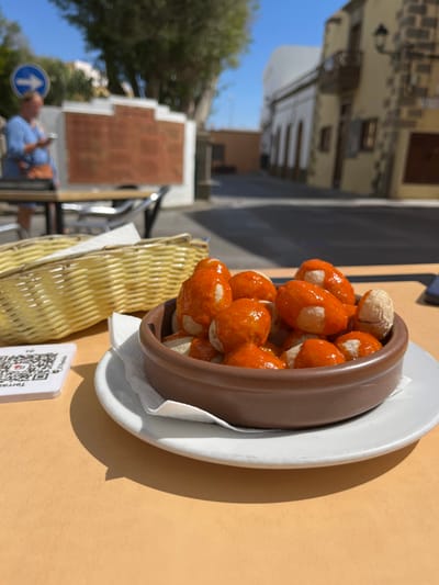 A brown ceramic bowl filled with round potatoes coated in a bright orange sauce rests on a white plate, which is placed on a light brown table. The bowl is partially obscured by a white napkin. A woven wicker basket is visible to the left of the bowl. To the left of the basket is a white square object with a black QR code and white text. The scene is an outdoor patio in the town of Agüimes, Spain, located at coordinates 27.907 latitude, -15.447 longitude. A blurred figure stands in the background, out of focus, wearing blue clothing.  A brick wall with a row of trees in front of it is visible in the distance. The sky is clear and blue.