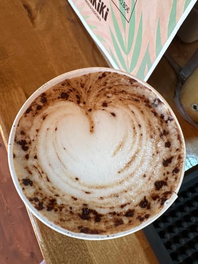 A close-up, overhead shot shows a heart shape made of foam and chocolate powder on top of a latte served in a white paper cup. The cup is positioned on a wooden table next to a pink menu with green leaves and text. The menu mentions "Kiki" and "SNACKS & COCKTAILS" with a price of "0.80€". In the background, a dark surface with raised circular bumps is visible, possibly a bar counter. The lighting is warm, suggesting an indoor setting. The image is a high-angle shot, focusing on the coffee art. The context suggests this scene is from a cafe or bar in Arroyo De La Miel-Benalmadena Costa, Spain.