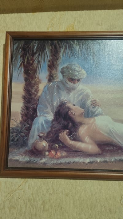 A framed painting depicts two figures in a desert setting. A person wearing a white turban and robes is seated, cradling a woman who is lying down. The woman has long, dark hair and is wearing a white garment. In the foreground, there is a small vase and some fruit. Palm trees are visible behind the figures. The painting is hanging on a textured yellow wall, with some light reflecting off the wall's surface. The overall impression is of a peaceful scene, possibly depicting a romantic or tender moment. The image does not provide any information related to the city of Dagomys, Russia.