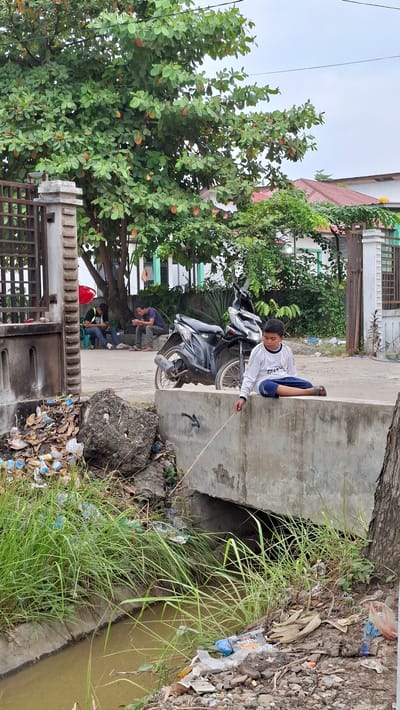 The image depicts an outdoor scene in Keudebayu, Indonesia, likely a roadside or urban drainage area. In the foreground, a young boy wearing a white long-sleeved shirt and blue shorts sits on the edge of a concrete structure, dangling a thin stick into a narrow, murky water channel below. The channel is lined with overgrown green grass and littered with various types of trash, including plastic bottles, wrappers, and debris.

Behind the boy, a black motorcycle is parked on a paved surface. Further in the background, two men are seated near a fence, appearing to be engaged with their mobile phones. The environment is characterized by a large tree with green leaves and some reddish-brown foliage, providing shade. Residential buildings with red-tiled roofs and white walls are visible in the distance. The overall scene suggests a somewhat neglected urban or semi-urban area, with a focus on the boy's solitary activity amidst the surrounding clutter.