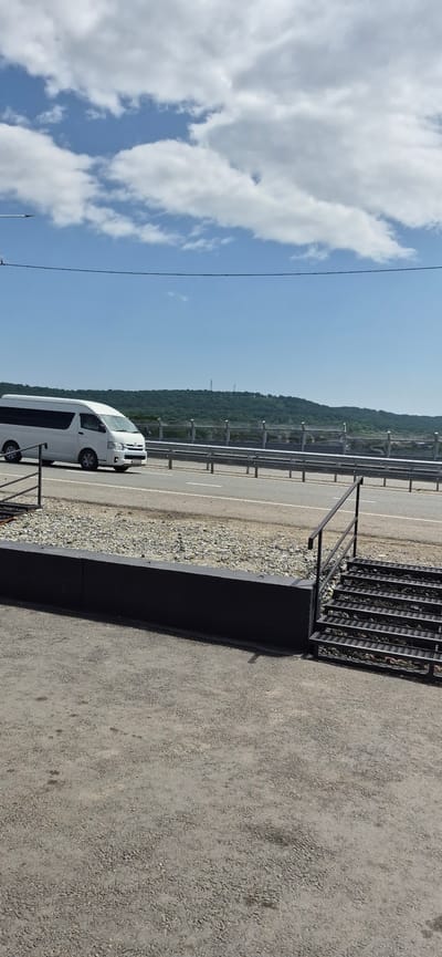 The image captures a scene on a sunny day, likely near the city of Taezhnyi, Russia, given the presence of a white minivan with what appears to be Russian license plates. The minivan is traveling on a highway, indicated by the clear lane markings and the guardrail separating the road from the adjacent embankment. In the foreground, a dark, rectangular concrete barrier runs horizontally, with a metal staircase featuring perforated steps attached to its right side. Beyond the highway, a lush, green, tree-covered hill rises, with cell towers visible at its summit. The sky above is a vibrant blue, punctuated by scattered white clouds. The overall environment suggests an outdoor setting on a well-maintained road.