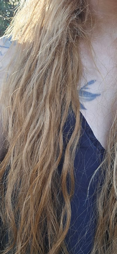 The image is a close-up, vertical shot of a person's head and upper torso, with a dark blue garment visible. Long, wavy, blonde hair dominates the frame, obscuring much of the face. A tattoo resembling stylized wings or tribal markings in a dark blue hue is visible on the person's cheekbone, partially hidden by the hair. The lighting suggests an outdoor setting, with sunlight catching the strands of hair and creating highlights. The background is blurred, with hints of greenery, possibly foliage. The exact location within Radovići, Montenegro, cannot be determined from the image alone.