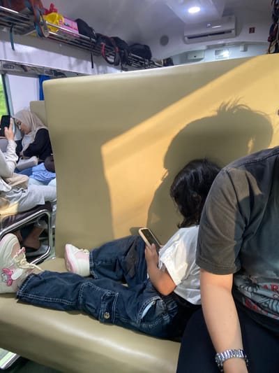 A young child sits on an upholstered seat in a vehicle, possibly a train or bus. The child is wearing a white shirt, blue jeans, and pink and white shoes. They are holding a smartphone. An adult figure is positioned above the child, partially obscuring them from view. Another seated passenger is visible near the floor. Luggage racks and an air conditioning unit are also visible within the train car. The location is Winong, Indonesia.