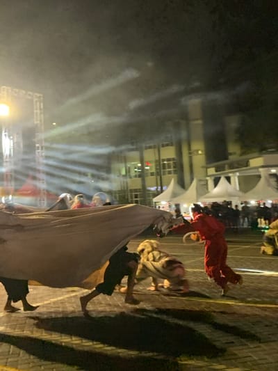 The image shows a nighttime street performance in Kecamatan Batu, Indonesia, a city located at the coordinates -7.867 latitude and 112.514 longitude. The performers are dressed in traditional costumes, with one person wearing a large, white cloth draped over their body, another wearing a black garment, and a third person wearing a red costume. The person in the white cloth is interacting with a person in a tiger costume by holding the tiger's tail. The person in the red costume is running away from the performers. The performance is taking place on a paved street illuminated by streetlights. The background features a building with large windows and multiple white tents. The air is filled with smoke from the performance.