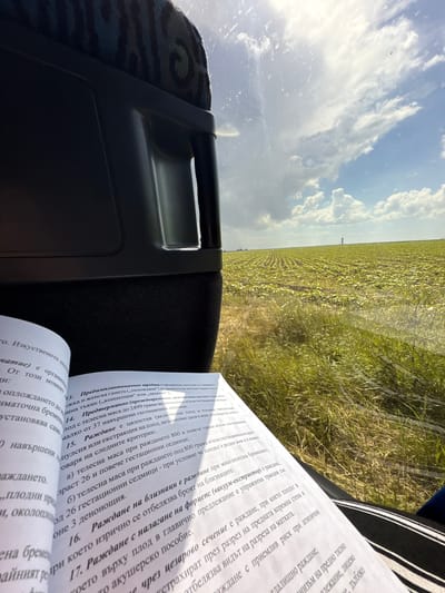 The image appears to be taken from inside a vehicle, possibly a bus or car, offering a view of a field and sky through the window. A book written in Bulgarian is visible on the right side of the frame, possibly on the seat next to the photographer. The location may be Byala Slatina, Bulgaria, considering the text in the book. The scene includes elements of transportation, reading, and a rural landscape. The book contains information on the subject of pregnancy.