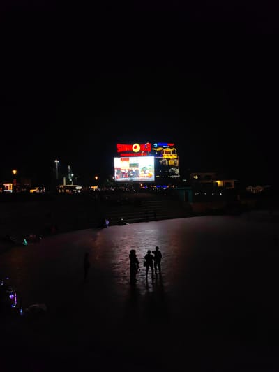 The image depicts a nighttime scene at a public plaza within the Sun World Fansipan Legend complex in Sa Pa, Vietnam. A large LED screen dominates the background, displaying colorful animated content and featuring prominent red and yellow signage for "SUNWORLD" and "FANSIPAN LEGEND." Below the screen, a crowd of indistinct figures is gathered on multiple tiers of steps and an elevated area, observing the display.
In the midground-left, a white car and a white bus or van are visible on a street, alongside streetlights. The foreground consists of a vast, dimly lit, paved surface. Five human figures are present in this foreground area, appearing as dark silhouettes against the faint reflections on the ground. Four figures are grouped closely together in the center-right, while a single figure stands further left. Near the isolated figure on the left, several objects on the ground emit subtle, colorful lights, primarily blue and green. The overall environment is a bustling public space characterized by artificial illumination against a dark sky.
