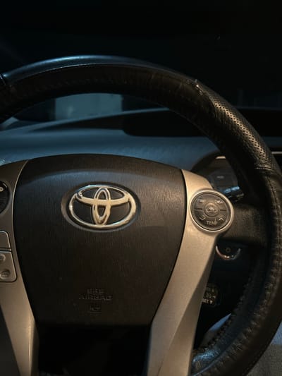 The image shows a close-up of a car's steering wheel. The steering wheel is black leather with visible stitching. A silver trim surrounds the center of the steering wheel, which features the Toyota logo. On the right side of the steering wheel, there are several buttons. One circular button cluster has labels for "TRIP," "DISP," a car icon, and "TEMP," along with up and down arrows. Another button with a power symbol is visible further down. On the left side of the steering wheel, there are buttons that appear to be for phone controls. The "SRS AIRBAG" text is embossed on the center airbag cover. The background is dark and out of focus, suggesting an interior shot of a vehicle at night. The location is specified as Gaidar, Moldova.