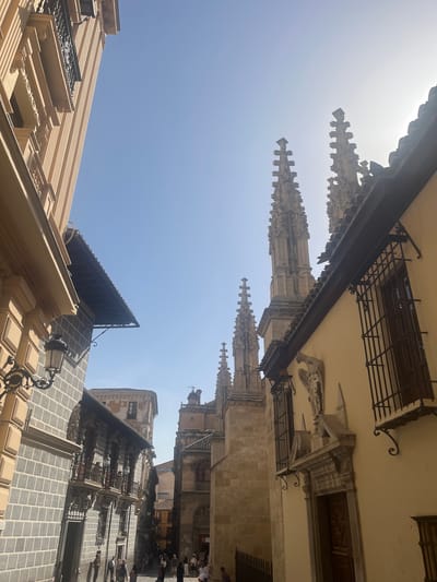 The image depicts a street scene in Granada, Spain, featuring a blend of architectural styles. On the left, a building with a light-colored, vertically striated facade is visible, adorned with decorative elements and a wrought-iron lamp. Adjacent to it is another building with a grey, tiled facade and ornate balconies.
Dominating the background are the spires of a Gothic cathedral or church, characterized by intricate stonework and pointed pinnacles. The sun is bright, casting shadows and creating a slightly hazy atmosphere. In the foreground and mid-ground, several people are walking along the cobblestone street, suggesting a bustling urban environment. The buildings on the right have a more classical aesthetic, with a pale yellow exterior, barred windows, and decorative architectural features above a doorway. The overall scene captures a historical and visually rich section of the city.