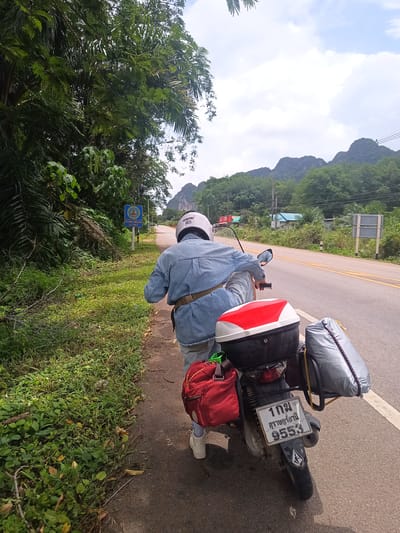 A person wearing a white helmet, denim shirt, and light-colored pants is shown from behind, walking a motorcycle on the side of a paved road in Khao Khram, Thailand. The motorcycle is heavily laden with a red backpack, a black top case with a red and white top, and a large grey bag. The motorcycle's license plate reads "1 กม สุราษฎร์ธานี 9553". The road has yellow dividing lines, and lush green vegetation grows along the roadside. A blue and white sign is visible near the vegetation. Mountains are visible in the background.