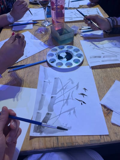 The image displays an overhead view of a painting activity taking place on a light-brown wooden table. At least three individuals are partially visible, indicated by their hands, each holding a paintbrush and actively painting on white paper sheets. The central and most prominent artwork features a monochromatic depiction of bamboo stalks and three dark, bird-like silhouettes. To the right, another paper shows practice strokes or elements.

Essential objects on the table include: a white circular paint palette with a prominent dark (likely black or dark green) paint mixture in its central well, flanked by smaller wells containing yellow, green, and blue pigments; multiple paintbrushes of varying sizes, some with blue handles; a clear square plastic container holding light blue water, presumably for rinsing brushes; and several graphite pencils. Beverages present are a wine glass with red liquid and a clear plastic bottle with a pink cap, labeled "MODEON." A small clear plastic bag containing purple, translucent objects and a stack of white paper napkins are also visible.

The scene captures individuals engaged in focused artistic creation, sharing common art supplies. The presence of the "MODEON" bottle, a specific Colombian brand of flavored water, indicates the location is an indoor establishment, likely an art studio or cafe, within Bogota, Capital District, Colombia.