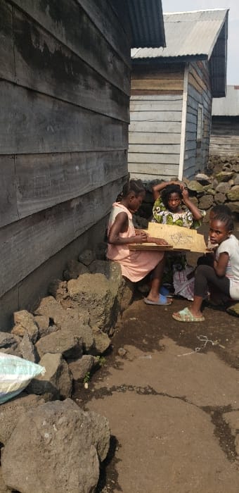 The image depicts an outdoor scene in Buzove, Democratic Republic of the Congo, featuring three individuals near two wooden-plank structures with corrugated metal roofs under clear daylight. In the foreground, the ground is composed of compacted dirt with visible cracks and numerous dark, irregularly shaped rocks.

From left to right, a young female in a light-colored dress sits on a pile of these rocks, holding a rectangular wooden board with visible markings on its surface. She appears engaged with the board, looking down. Her feet are shod in blue slide sandals. Behind her, an older female or youth, dressed in a patterned garment featuring green, white, and yellow hues, sits in a slightly elevated position, with her hands near her head. To the right, another young female, wearing a white short-sleeved top and dark pants, sits directly on the dirt ground, looking towards the viewer. She wears patterned slide sandals.

On the far left, a portion of a white and blue patterned sack is visible on the ground. The building immediately to the left is constructed of weathered, horizontal wooden planks, with a corrugated metal roof. Another similar wooden building with a corrugated metal roof is visible behind and to the right of the individuals. The overall environment suggests a residential or community area.