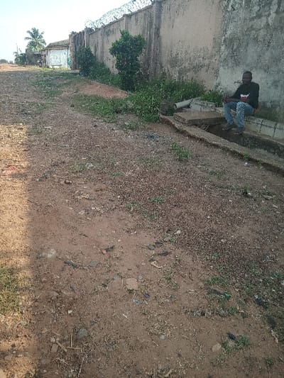 The image shows an open dirt area in Ota, Nigeria (latitude: 6.689, longitude: 3.176). The dirt is mostly bare, but there are patches of grass growing in it. A concrete curb and a concrete retaining wall run along the right side of the image. A man is sitting on the retaining wall, facing the camera. He is wearing a dark-colored shirt, blue jeans, and sandals. He is holding a white object in his right hand. The top of a building is visible in the background.