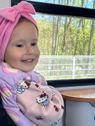 A young child is depicted in a close-up, eye-level shot, smiling broadly at the camera. The child is wearing a pink ribbed headband with a large bow on the left side of their head. They are dressed in a light purple quilted jacket featuring a pattern of rainbows and apples, layered over or next to a pink top or blanket adorned with white Hello Kitty characters and red cherries.
The child's expression is joyful, with a wide, open-mouthed smile revealing several lower and upper teeth. Their dark eyes are bright and engaging. The setting appears to be indoors, next to a large window with a dark frame. Outside the window, vibrant green foliage from trees is visible, suggesting a natural outdoor environment. A white, grid-like fence or railing is also discernible through the window. Further in the background, a warm-toned wooden element and a light grey surface are present. The lighting is bright and natural, indicating daytime. The child's warm clothing, including a quilted jacket, suggests the weather might be cool. The image was taken in Ustroń, Poland.