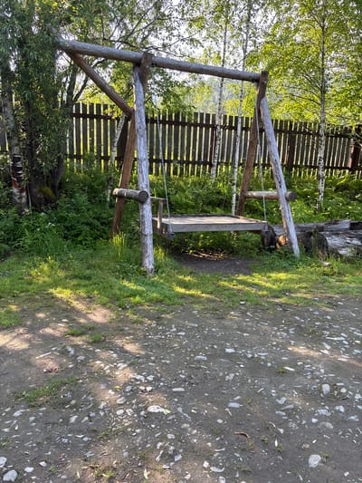 The image depicts a rustic wooden swing set set in a natural, outdoor environment. The swing set is constructed from rough-hewn logs and branches, with a wooden plank seat suspended by chains. It is positioned in a grassy area with a dirt path in the foreground, marked by scattered stones and patches of green vegetation. Behind the swing set, a wooden picket fence runs horizontally across the frame, separating the yard from a densely wooded area. The trees in the background are predominantly birch trees with their characteristic white bark, interspersed with other deciduous trees displaying lush green foliage. Sunlight filters through the leaves, casting dappled shadows on the ground. The overall impression is one of a tranquil, rural setting, possibly a backyard or a park in a natural area. The location is identified as Bol'shaya Rechka, Russia, suggesting a region with a prevalence of birch forests and a natural, somewhat wild landscape. There are no people or animals visible in the image.