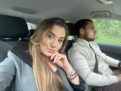 The image depicts two people inside a car. A woman with long blonde hair, wearing a gray jacket, is in the foreground, facing the camera. She has a bracelet and a watch on her left wrist and pink-tipped nails. A man with a beard and short dark hair, wearing a light-colored jacket, is visible in the background, looking out the window. Both appear to be wearing seatbelts. Outside the window, green foliage is visible, suggesting a natural environment. The car's interior includes a gray dashboard and black seats. The location, according to the request, is Krupnik, Bulgaria, implying the photo was taken there.