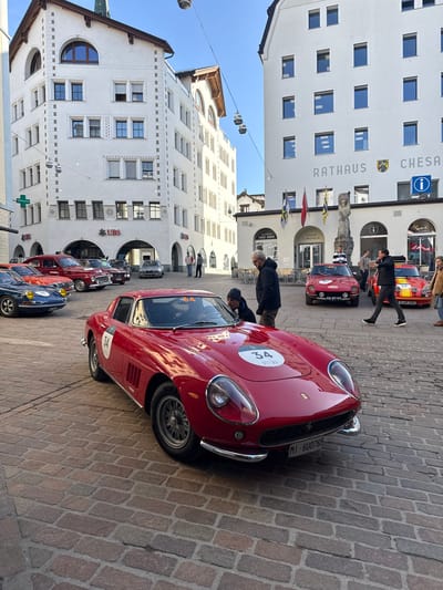The image presents a daytime scene in the cobblestone-paved square of Sankt Moritz, Switzerland. The central focus is a bright red vintage Ferrari, identified by '34' white circular decals on its hood and driver's side fender, parked at an angle in the foreground. The car displays an Italian license plate: MI-6U0765. Two men in dark jackets are positioned near this Ferrari; one stands looking into the driver's side, while the other leans into the passenger compartment.

In the mid-ground, a collection of other vintage automobiles is visible. To the left, a blue vintage car with yellow fog lights, marked with the number '30', is partially seen. Further back, a red vintage sedan is parked alongside other classic vehicles. Towards the right, a red vintage car with '40' on its hood and license plate AR-87-95 is parked, and next to it, a yellow and red striped vintage car with '39' on its hood and license plate S-WP 20H.

Several individuals populate the scene. A man in a black jacket walks from right to left across the square, looking towards the parked cars. Other people stand near the vehicles or by the buildings.

The square is flanked by multi-story white buildings under a clear blue sky. On the right, the "RATHAUS CHESA CUMUNELA" (Town Hall of St. Moritz) features prominent lettering, a crest, multiple rectangular windows, and a stone statue in front of its entrance, beneath a display of flags. A sign for "TAKE AWAY" and an information sign (blue circle with 'i') are also visible. On the left, another white building with varied window designs and a distinctive arched window near its peak houses a "UBS" bank branch and an "AMAVITA" pharmacy, indicated by a green cross sign. Overhead wires with suspended light fixtures are visible against the sky. The ground throughout the square is covered in reddish-brown cobblestones.