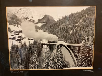The image is a black and white photograph of a steam train crossing a snow-covered viaduct in a mountainous, forested region. The train is emitting a large plume of white steam. Below the viaduct, a village with small, dark buildings is nestled amidst snow-laden evergreen trees. The background features towering, snow-capped mountains. The bottom left corner of the image has handwritten text that reads "28. JANUAR 1968". The bottom right corner has handwritten text that reads "FOTO HOHENBERGER, AROSA". The photograph is framed by a dark mat and a light brown border. The location is not identified as Ленинское, Russia, but rather Arosa, Switzerland based on the photographer's credit.