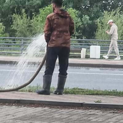 A person in black pants and a brown jacket, wearing black rubber boots, is holding a hose that is spraying water onto a sidewalk. The water is creating a mist. The person's face is partially visible, and they appear to be looking down at the hose. In the background, another person in light-colored clothing and a hat is leaning over, possibly observing the first person or the surroundings. The scene appears to be outdoors, with trees and a road visible. The weather seems to be overcast, as there are no strong shadows. The ground is paved with interlocking bricks and concrete. The setting could be a street or a park. The time of day is indeterminate. There is no visible text in the image. The location is Aktobe, Kazakhstan.