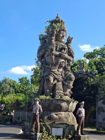 The image captures a monumental stone statue of a multi-armed deity, weathered and partially covered in moss, standing prominently in Banjarangkan, Indonesia. The statue is positioned on a lotus-shaped pedestal, with two life-sized statues of what appear to be police officers flanking it, one on each side. These figures are dressed in traditional police uniforms, complete with hats, shirts, belts, and trousers, and are positioned with their hands behind their backs. The background features lush green trees and a clear blue sky with scattered clouds. An arched gateway, also made of stone and adorned with traditional carvings, is visible behind the main statue. The scene is set outdoors, likely in a public space or at the entrance to a significant location, with a road visible in the foreground.