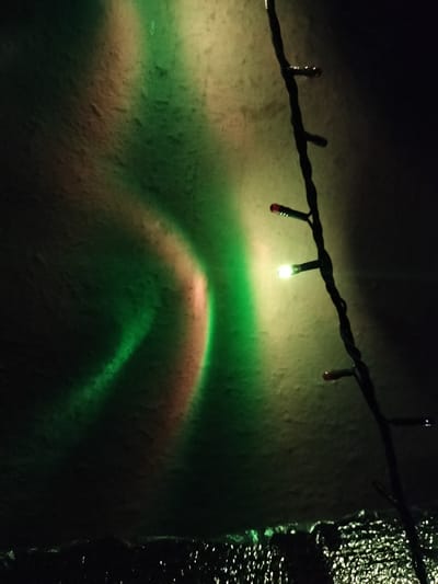 This is a nighttime photograph showing a string of Christmas lights casting green and pinkish light on a textured, light-colored surface. The string of lights is dark and runs diagonally from the upper right to the middle of the frame, with several individual LEDs visible. One LED in the middle is lit with a bright white or yellow light, and there are also red LEDs visible at the top and bottom of the visible string. The light on the surface creates blurred, abstract patterns of green and a reddish-pink hue, suggesting a colorful aurora-like effect. The bottom portion of the image is dark and appears to show some sort of reflective or textured material. The image does not contain any identifiable people or specific landmarks that would place it in Kudashivka, Ukraine.