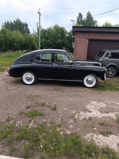This media file shows a black vintage car parked on a gravel driveway in front of a brick garage. The car is a sedan with a sloping roofline and chrome accents. It has white-wall tires with chrome hubcaps. A dark gray SUV is parked to the right of the vintage car, partially obscured by it.
The setting appears to be rural or semi-rural, with trees and greenery visible in the background. A utility pole with electrical wires is also present. The ground is covered in gravel and patches of grass and wildflowers. The sky is overcast, suggesting a cloudy or partly cloudy day. The overall impression is that of a casual outdoor scene, possibly a residential area or a storage lot. There is no discernible activity taking place; the vehicles are stationary.
Visible text includes a red and white emblem on the side of the vintage car, reading "GAZ-21". The SUV has "4x4" lettering on its rear. The image does not provide clues about the time of day or convey any specific emotions. The location context is provided as Tolbazy, Russia.