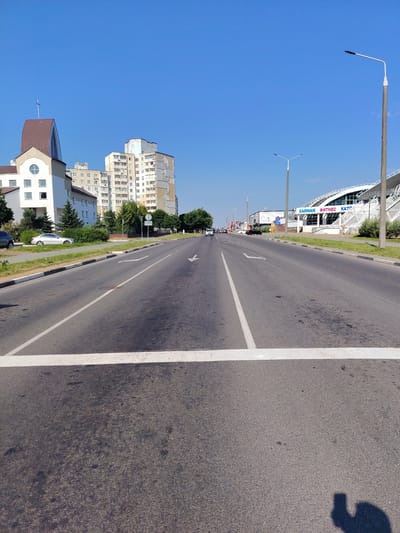 The image shows a wide, straight road in Salihorsk, Belarus. On the left side of the road, there is a modern building with a distinctive curved roof topped by a cross, resembling a church or a religious building, next to several multi-story residential buildings. On the right side of the road, there is a large, white, modern building with a curved roof structure, labeled "ХАММАМ ФИТНЕС КАТС". The sky is clear and blue. The road has lane markings and a pedestrian crossing marked by a thick white line in the foreground. There are a few cars visible on the road and on the side of the road. The overall scene depicts a typical urban environment on a sunny day.