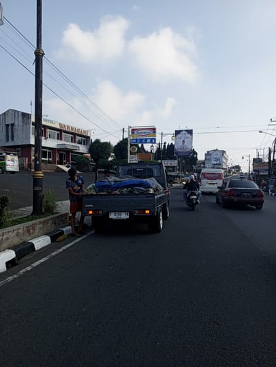The image captures a bustling daytime street scene in a city, identifiable by the presence of an Indomaret convenience store sign and Indonesian license plate formats on visible vehicles. On the left, a multi-story building with "HOTEL WARNASARI" prominently displayed on its facade stands alongside the road. In the foreground, a tall, dark utility pole with overhead wires extends upwards.
A dark gray Suzuki Carry pickup truck is parked or stopped on the left side of the road, close to a curb painted in black and white stripes. Its tailgate is open, revealing a cargo bed filled with numerous items, possibly produce like large fruits or vegetables, partially covered by a blue tarp. A young male in a blue patterned shirt and brown shorts stands next to the open tailgate, appearing to tend to the cargo. He is wearing flip-flops. Another person is visible inside the cab of the pickup. The truck's license plate, "F 8180 YN," is clearly visible. A coil of yellow material rests on the curb near the young man's feet.
Further down the road, traffic is moving. A motorcyclist in a white helmet and dark jacket is riding on the asphalt road, positioned behind the pickup truck. A white passenger van with "Rhis.id" printed on its side is visible further ahead, followed by a black sedan with the license plate "F 1347 YL." In the background, several billboards are visible; one prominently features "IDUL ADHA" with an image of a man, and another advertises "SINAR KAPA." The sky above is clear blue with scattered white clouds. The overall environment suggests an urban or semi-urban commercial area with various businesses lining the street.