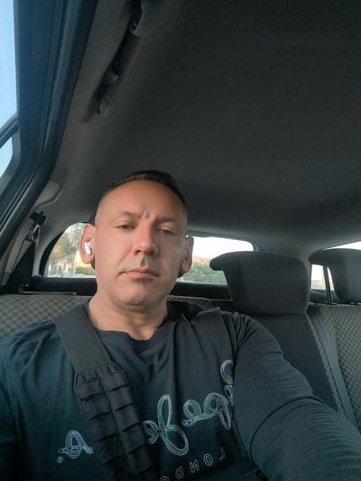 A man is sitting in the driver's seat of a car, looking at the camera. He is wearing a dark t-shirt and has white earbuds in his ears. The interior of the car is visible, including the headrests and seats, which are upholstered in a checkered pattern. Outside the car, a few trees and buildings are visible, suggesting an urban or suburban setting. The sky appears clear and bright. The location is the Municipal Unit of Acharnes, Greece.