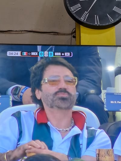 The image shows a man wearing sunglasses and a white polo shirt with green and red accents, sitting in a stadium. A scoreboard on a television screen displays "34:24" and "MEX 0 0 KOR", indicating a soccer match between Mexico and South Korea. The man has a silver chain around his neck. In the background, a large clock with Roman numerals is visible on a yellow wall. The scene appears to be in the city of Bireuen, Indonesia.
