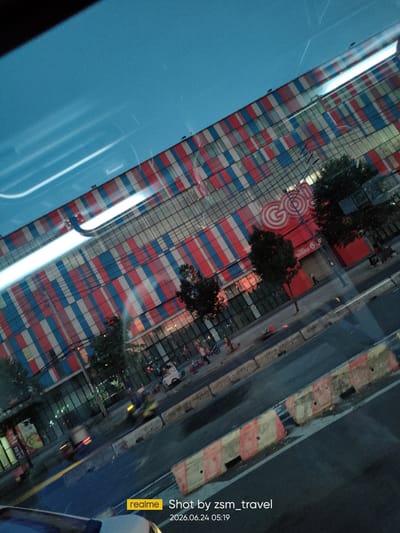 The image shows a street scene in Thuận An, Vietnam, likely captured from a vehicle. In the background, a large building with a distinctive facade of red, white, and blue rectangular patterns dominates the view. A prominent red sign with a white circular logo reading "GOON" is visible on the building's lower level. A street runs in front of the building, with a sidewalk lined with trees. Several vehicles, including motorcycles and a small white truck, are present on the street, suggesting ongoing traffic. Some people can be seen on the sidewalk. The sky above is a clear blue. The foreground of the image is partially obscured by the interior of a vehicle, with a blurred dashboard and reflections visible. Traffic barriers, with orange and white markings, are positioned along the edge of the road. A timestamp at the bottom right corner indicates the image was taken on June 24, 2026, at 05:19. The overall impression is of an urban environment during daylight hours.