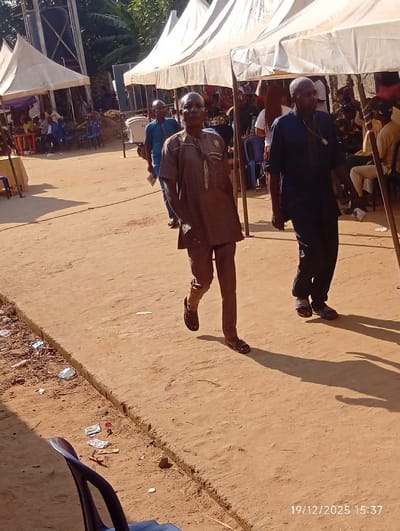 Under white tents, three men walk on a dusty, dirt road. The foreground man, dressed in brown, leads the group. Behind him, a man in blue follows. Tables and chairs are set up under the tents, where people are seated. Litter lines the ground. A date stamp reads 19/12/2025.