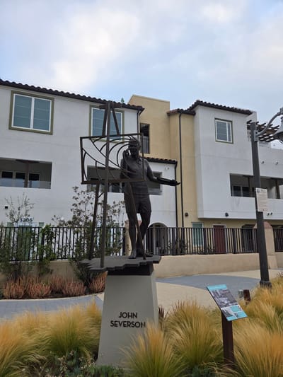The image shows a statue of a man, likely John Severson, in Dana Point, California. The statue depicts the man in a dynamic pose, with one arm raised as if reaching for something. He is dressed in athletic attire, with shorts and a shirt. The statue is made of dark metal and is placed on a pedestal that has "JOHN SEVERSON" inscribed on it.

The setting is an outdoor urban environment, possibly a park or a public plaza. There are modern buildings in the background, featuring white and yellow facades and windows. A metal fence is visible, separating the area with the statue from the buildings. In the foreground and to the right, there are tall, golden-colored ornamental grasses and some succulents. A sign with text and images is also visible near the grasses.

The sky is overcast, suggesting it might be a cloudy day, or perhaps early morning or late afternoon. The overall mood of the image is serene and contemplative, focusing on the statue as a tribute. The urban architecture contrasts with the natural elements of the plants. The inscription on the pedestal clearly identifies the subject of the statue.