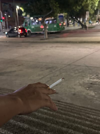 An outdoor, ground-level, medium shot captures a person's hand holding a lit cigarette in the foreground, with a city street scene in the background. The hand is dark-skinned, with the index finger and thumb holding the cigarette, which has an ember at the tip. The background is blurred, showing a street with a dark-colored car on the left, its brake lights illuminated, and a green bus further back. A traffic light with a red light is visible on the left side of the street. Trees and streetlights are also present in the background. The ground in the foreground consists of concrete steps, with the background ground appearing to be asphalt or pavement. The image suggests an urban environment, possibly in Perímetro Urbano Barranquilla, Colombia, during the evening or night due to the artificial lighting and illuminated vehicle lights.