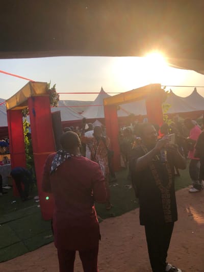 This image depicts an outdoor social gathering in the city of Amanuke, Nigeria, at what appears to be late afternoon or sunset. The scene includes multiple individuals and temporary structures under a bright, low sun positioned in the upper right, casting a warm glow.
In the foreground, two male subjects are prominent. On the left, with his back to the camera, is a man wearing a maroon long-sleeved tunic and matching trousers. A black and white patterned bandana or scarf is draped over his left shoulder. To the right, another man, facing towards the left of the frame, is holding a white smartphone horizontally, seemingly recording or photographing the event. He wears a black tunic with a distinctive gold-colored geometric pattern resembling a Greek key or chain down the front, dark trousers, and light-colored sneakers.
The ground consists of a reddish-brown sandy or dirt area interspersed with sections of green artificial turf or matting. In the mid-ground, a decorative archway structure, primarily red with gold trim and some green foliage accents, frames a view into the larger event space. Red ribbons or ropes are visible stretching between structures. Behind the archway, numerous other attendees are present, some engaged in conversation or movement; one man in a patterned orange and brown tunic has his right arm raised. Several white, peak-roofed event tents are visible in the background, extending towards the horizon.