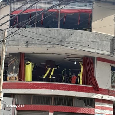 The image shows a building in Nuseirat, Palestinian Territories. The building appears to house a gym, as evidenced by the "EMPIRE GYM" sign. Gym equipment is visible inside. The building's exterior is a mix of concrete and red and white striped features. Electrical wires are strung across the foreground.