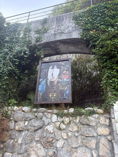 A framed painting depicting two figures, possibly mythological or allegorical, is displayed against a stone wall under a concrete bridge. The painting is tilted and leaning against a cluster of rocks and vegetation. The bridge, covered in dense green ivy, is a prominent feature of the environment. A metal railing runs along the top of the bridge. Behind the painting, a dark, narrow staircase with a black metal railing leads upwards into dense foliage. To the right of the painting, a metal plaque with Cyrillic writing is affixed to a grey stone structure, possibly indicating a historical or cultural site. The overall scene suggests an outdoor location in a somewhat overgrown, natural setting, likely within the city of New Athos, Abkhazia.