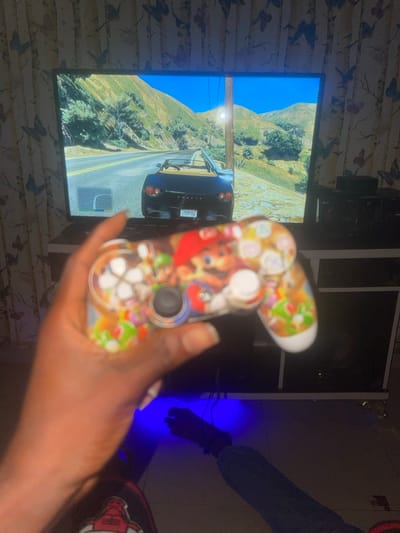A person holds a custom-designed game controller, featuring cartoon character graphics, in front of a large screen displaying a video game scene. The game shows a black car driving on a road amidst mountainous terrain under a clear blue sky. The background includes furniture and walls with patterned wallpaper. A separate figure partially visible has dark clothing and gloves. Blue lighting emanates from behind the furniture.
